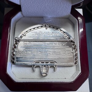 May include: A silver brooch featuring engraved hieroglyphs and the name "Sami Makhlouf". The rectangular brooch has a curved top and a decorative pin. It is displayed in a burgundy presentation box.