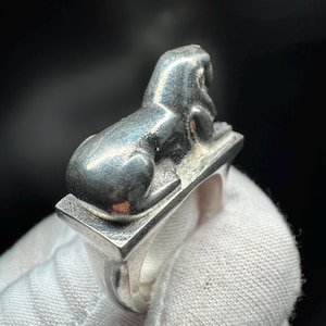 May include: A silver ring with a detailed sphinx design. The sphinx is seated on a rectangular base, with the ring band below. The ring has a polished finish and a vintage aesthetic, reflecting a classic style.