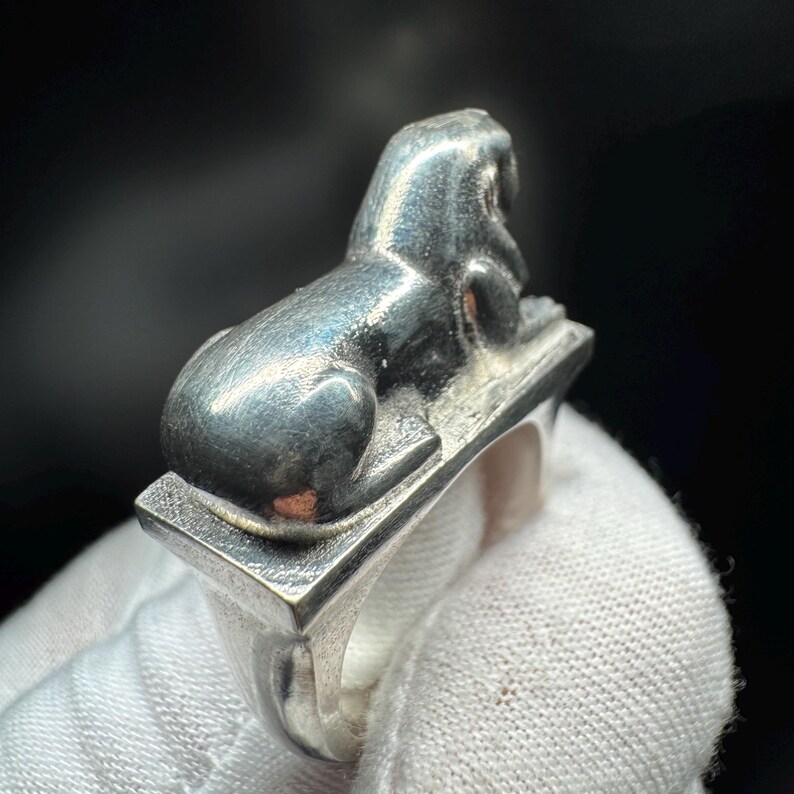 May include: A silver ring with a detailed sphinx design. The sphinx is seated on a rectangular base, with the ring band below. The ring has a polished finish and a vintage aesthetic, reflecting a classic style.