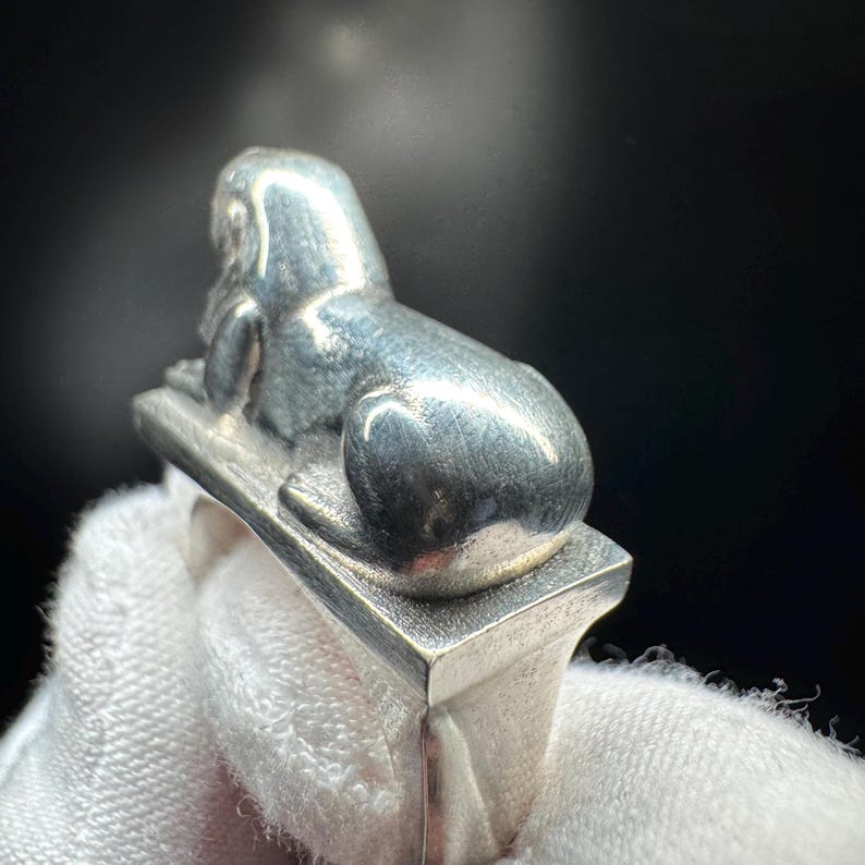 May include: A silver ring with a sphinx design. The sphinx is in a reclining position on a rectangular base. The ring has a polished finish and detailed design. The ring is likely made of sterling silver. The ring is a piece of jewellery.