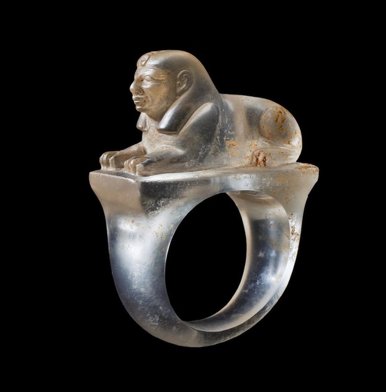 May include: A clear ring with a sphinx sculpture. The sphinx, with a human head and lion's body, sits on a rectangular base. The ring's band is smooth and rounded. The ring is made of a translucent material.