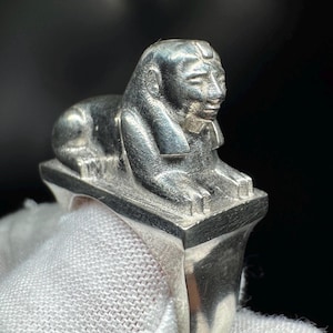 May include: A silver ring with a detailed sphinx sculpture. The sphinx is in a reclining position on a rectangular base, with detailed facial features. The ring's band is smooth and polished. The ring is a piece of jewellery.