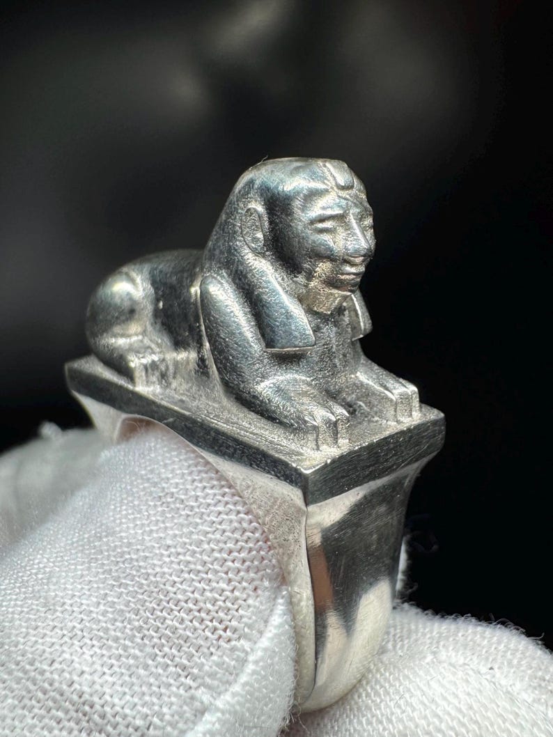 May include: A silver ring with a detailed sphinx sculpture. The sphinx is in a reclining position on a rectangular base, with detailed facial features. The ring's band is smooth and polished. The ring is a piece of jewellery.
