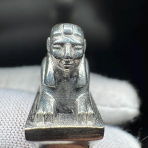 May include: A silver-coloured miniature sphinx sculpture. The sphinx is shown seated, with a human head and a lion's body. The sculpture has detailed features, highlighting the facial details and the overall form of the mythical creature.