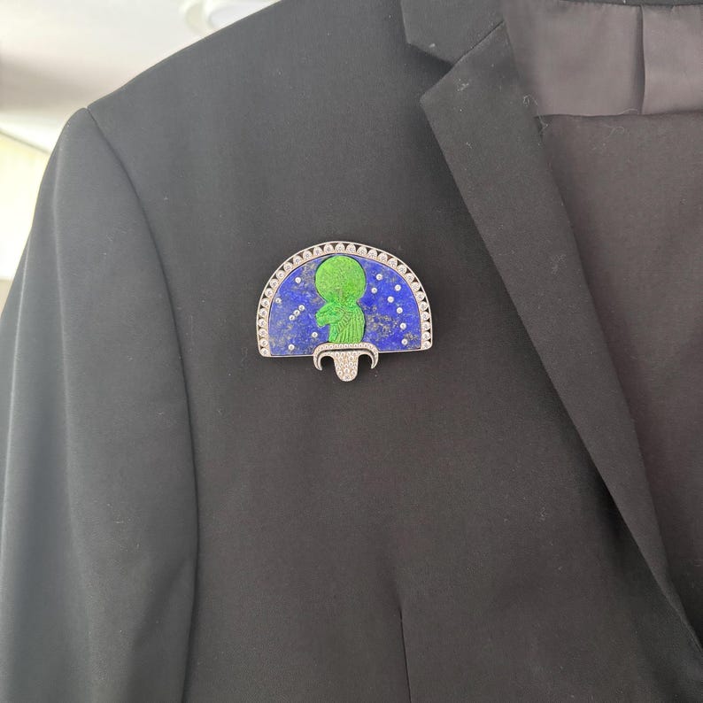 May include: A decorative brooch with a green figure on a blue background with white dots, set in a silver-toned frame. The brooch is attached to a black blazer, highlighting its design and craftsmanship. The brooch has a unique, artistic design.