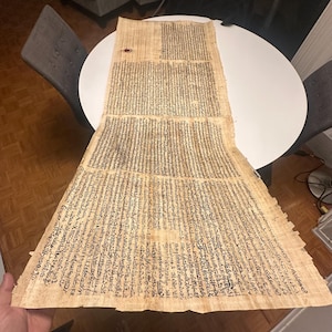 May include: A large, rectangular papyrus scroll with rows of black text. The papyrus is a light tan color, with a few darker spots. The scroll is partially unrolled on a white table, with the edges slightly curled.