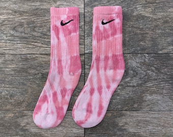 Puede incluir: Un par de calcetines de color rosa tie-dye con un logotipo de Nike negro en forma de swoosh en cada calcetín.