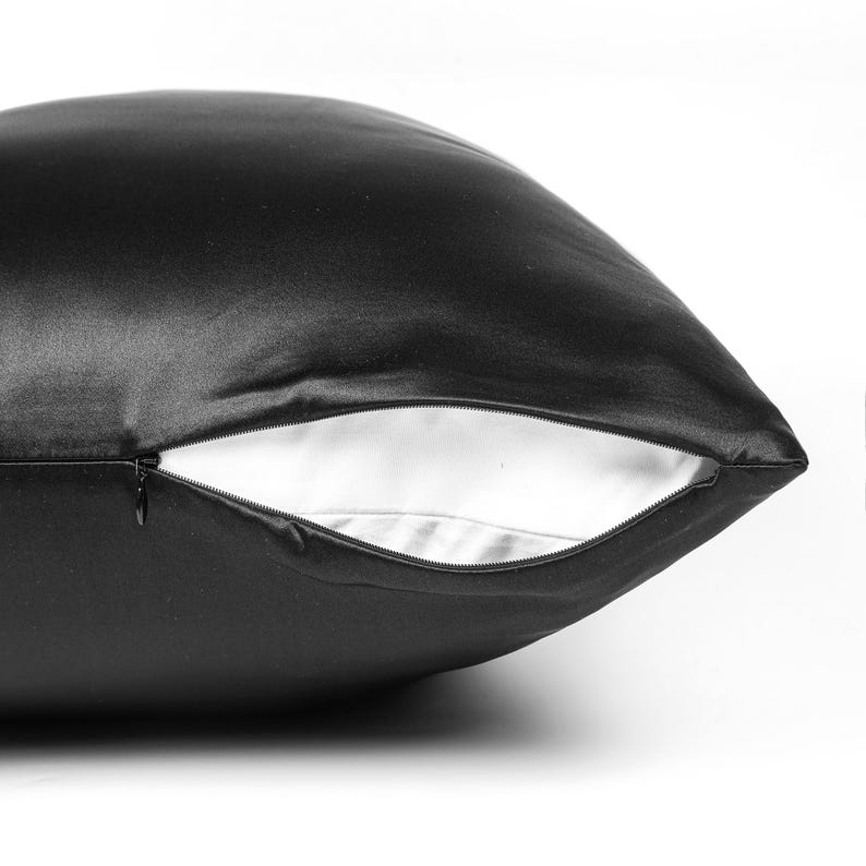 May include: A black pillowcase with a smooth, satin-like finish and a hidden zipper closure. The zipper is partially open, revealing a plain white interior lining. The fabric has a subtle sheen that reflects light across its curved surface.