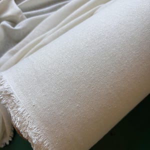 May include: A rolled length of off-white fabric, showing a textured weave and a fringed edge. The fabric is tightly wound, displaying its texture and colour. The material appears to be a natural fibre, possibly linen or cotton.