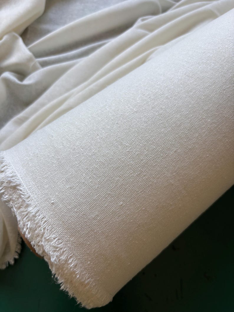 May include: A rolled length of off-white fabric, showing a textured weave and a fringed edge. The fabric is tightly wound, displaying its texture and colour. The material appears to be a natural fibre, possibly linen or cotton.