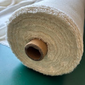 May include: A roll of cream-coloured fabric with a textured, fluffy edge. The fabric is wound around a brown cardboard core. The material appears to be a natural fibre, possibly cotton or linen, and is suitable for various textile projects.