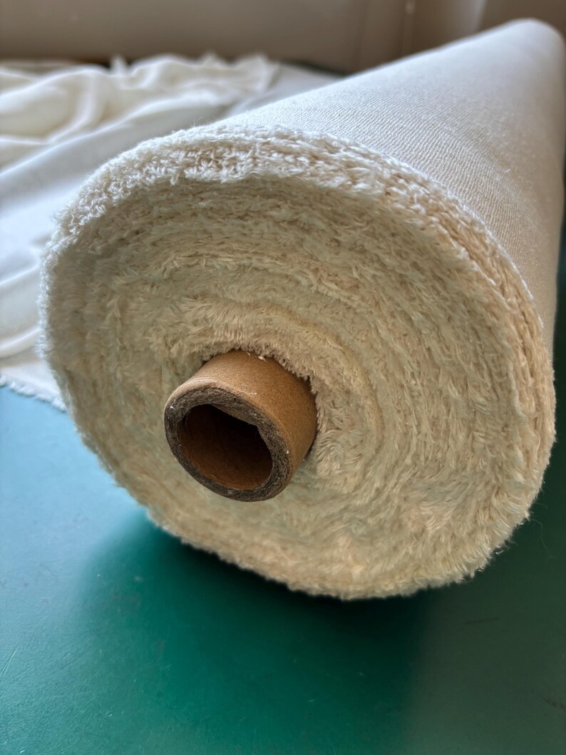 May include: A roll of cream-coloured fabric with a textured, fluffy edge. The fabric is wound around a brown cardboard core. The material appears to be a natural fibre, possibly cotton or linen, and is suitable for various textile projects.