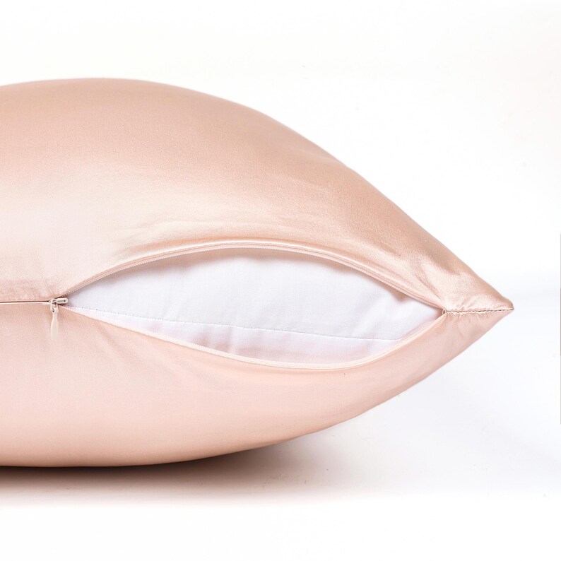 May include: A smooth, light pink pillowcase with a subtle sheen, featuring a partially open side seam that reveals a plain white pillow insert inside. The fabric has a soft, reflective texture and a concealed zipper closure along the edge.