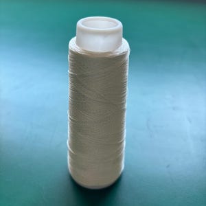 Könnte beinhalten: Eine Spule aus cremefarbenem Faden mit einem weißen Kunststoffaufsatz. Der Faden ist eng um die Spule gewickelt und bildet eine zylindrische Form. Der Hintergrund ist einfarbig grün.