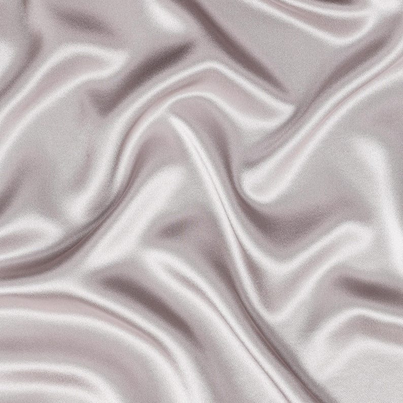 May include: A smooth, light mauve fabric with a lustrous, reflective surface. The material is draped in soft, flowing folds that create deep shadows and bright highlights across the surface, emphasizing the silky texture and consistent color.