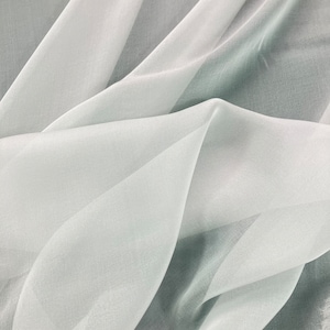 May include: A close-up of a sheer, light gray fabric with a soft, flowing texture. The fabric is draped and folded, creating a delicate and airy appearance.