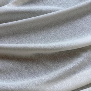 May include: A close-up shot of a light grey fabric, showing soft folds and a subtle texture. The material appears to be lightweight, possibly linen or a similar fabric, with a slightly translucent quality. The overall effect is one of gentle movement.