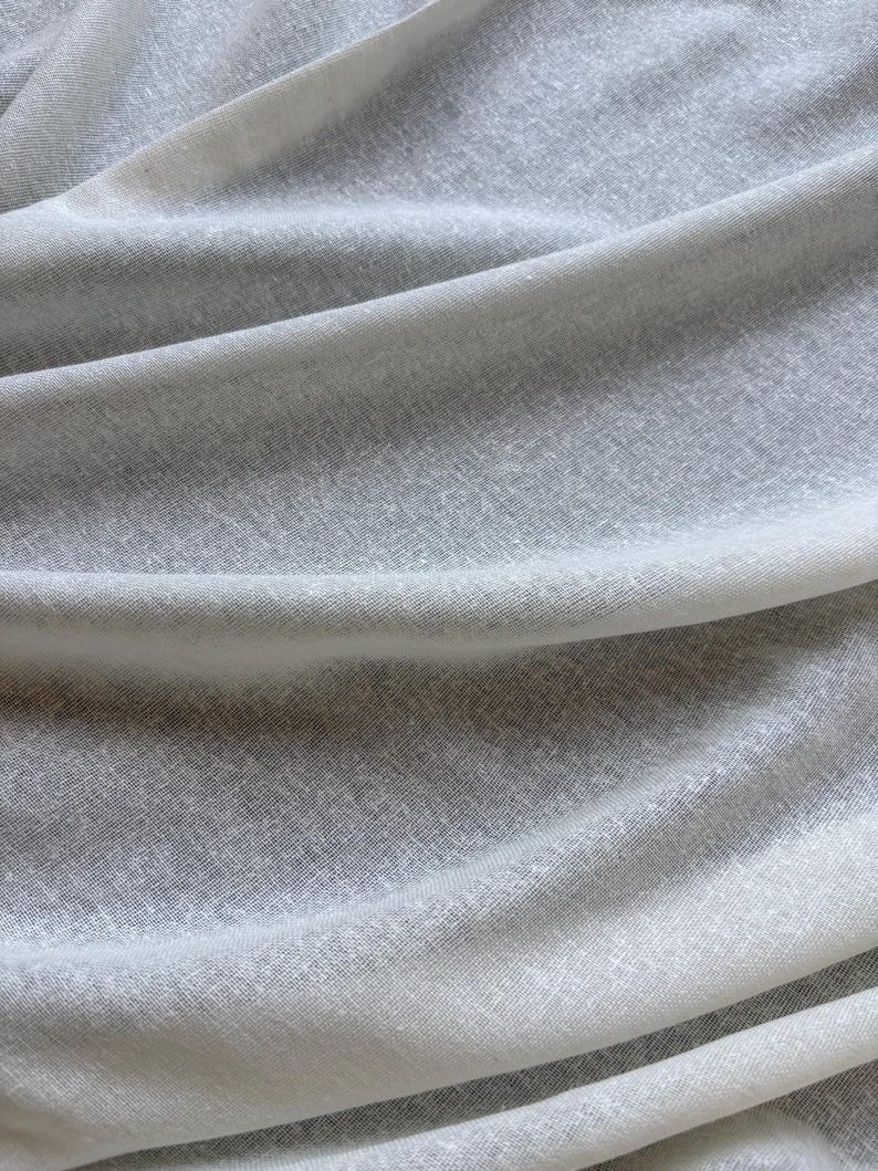 May include: A close-up shot of a light grey fabric, showing soft folds and a subtle texture. The material appears to be lightweight, possibly linen or a similar fabric, with a slightly translucent quality. The overall effect is one of gentle movement.