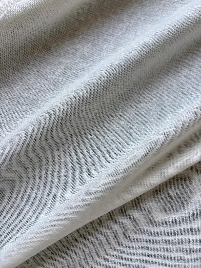 May include: Close-up shot of a light grey fabric with a textured, woven appearance. The fabric is draped, showing folds and subtle variations in tone. The material appears soft and lightweight, suitable for clothing or home decor.