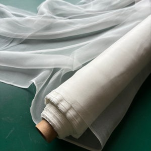 Puede incluir: Un rollo de tela blanca transparente, probablemente tul u organza, se muestra sobre una superficie verde. La tela es suave y delicada, con un ligero brillo.