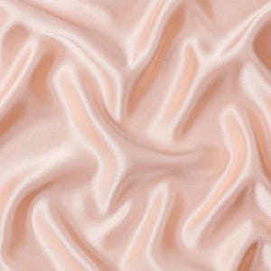 May include: A light blush pink fabric with a smooth, lustrous surface and a soft sheen. The material is draped in deep, flowing folds that create a series of highlights and shadows across the surface, emphasizing the fluid texture of the textile.