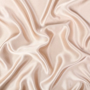 May include: A piece of light beige fabric with a smooth, lustrous finish and a soft sheen. The material is draped in deep, flowing folds that create a series of highlights and shadows across the surface, emphasizing the fluid texture of the cloth.