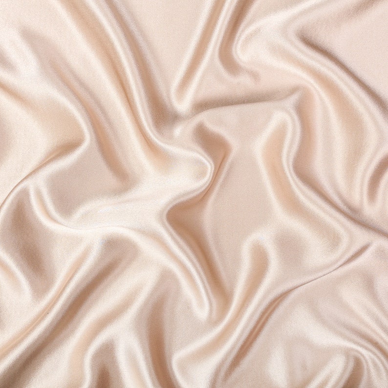 May include: A piece of light beige fabric with a smooth, lustrous finish and a soft sheen. The material is draped in deep, flowing folds that create a series of highlights and shadows across the surface, emphasizing the fluid texture of the cloth.