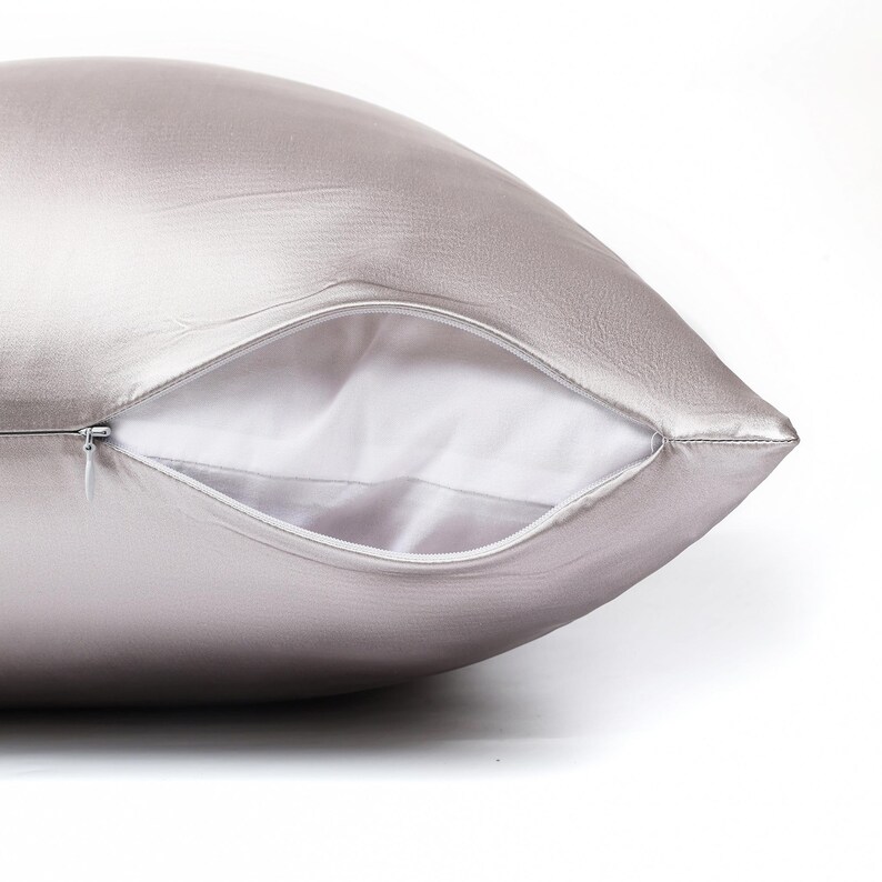May include: A smooth, light silver-colored pillowcase with a subtle sheen. The pillowcase features a concealed zipper closure along the side, which is partially open to reveal a plain white interior lining against the metallic silver fabric exterior.