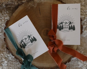 May include: Two wedding vow books with mountain and sunburst illustrations, tied with teal and rust-colored ribbons. The books are on a wooden slice, with the text "her vows" and "his vows" in elegant script.