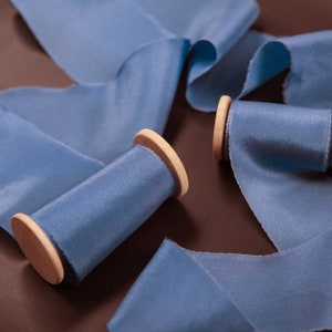 May include: Two spools of blue satin ribbon with wooden spools. The ribbon is arranged in a decorative way on a brown surface.