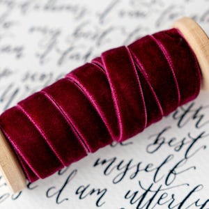Könnte beinhalten: Eine Spule mit tief burgunderfarbenem Samtband. Das Band ist eng um eine hellfarbene Holzspule gewickelt. Der Hintergrund zeigt eine elegante Schreibschrift in schwarzer Tinte, die dem Bild einen Hauch von Raffinesse verleiht.