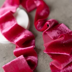 May include: Close-up of vibrant fuchsia velvet ribbon, artfully arranged on a neutral gray surface. The ribbon's texture is soft and plush, with a slight sheen. The ribbon is in a decorative arrangement, perfect for crafts and embellishments.