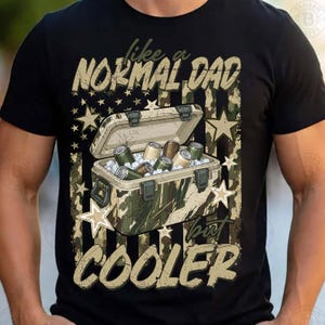Puede incluir: Una camiseta negra con un gráfico de una nevera portátil con estampado de camuflaje llena de latas de bebida. El diseño incluye una bandera estadounidense de camuflaje de fondo con estrellas y el texto "like a NORMAL DAD but COOLER" en una fuente desgastada.