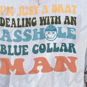Op de afbeelding: Lichtgrijs t-shirt met de tekst "I'M JUST A BRAT DEALING WITH AN ASSHOLE BLUE COLLAR MAN" in oranje, groen en blauw. Een smiley met een blauwe helm is in het ontwerp opgenomen.