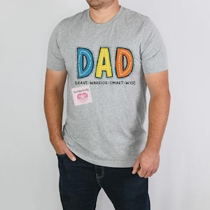 May include: A gray t-shirt with the word "DAD" in colorful, outlined letters. Below the word "DAD" is the text "BRAVE-WARRIOR-SMART-WISE". A small pink heart-shaped sticker is affixed to the shirt.