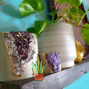 May include: Two white ceramic planters with gold accents and crystal accents. One planter has purple crystals and the other has clear crystals with gold and purple accents. The planters are on a wooden shelf.