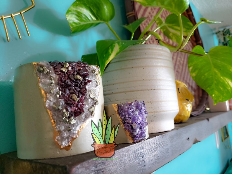 May include: Two white ceramic planters with gold accents and crystal accents. One planter has purple crystals and the other has clear crystals with gold and purple accents. The planters are on a wooden shelf.