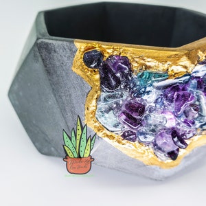 May include: A gray, geometric planter with a gold-leafed edge and a cluster of colorful, gem-like stones. The planter has a dark interior and a small, illustrated logo of a potted succulent.