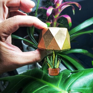 Puede incluir: Un macetero de hormig&oacute;n geom&eacute;trico dorado con un dise&ntilde;o angular y afilado. El macetero se sostiene en una mano sobre un fondo de plantas verdes y moradas.