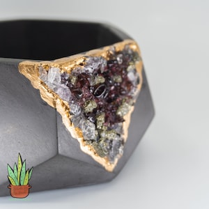 May include: A black geometric concrete planter with a gold-leafed, crystal-filled design. The planter is filled with a small green succulent plant.