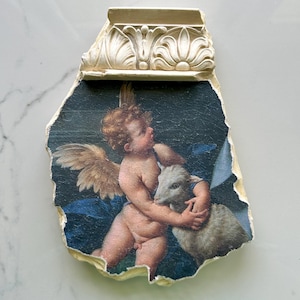 May include: A cherub with golden wings holds a white lamb in its arms. The cherub is painted in a realistic style with a blue background. The image is framed in a white plaster molding.