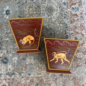 May include: Two decorative wooden boxes with a red background and yellow trim. Each box features a hand-painted image of a monkey in a jungle setting.