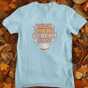 Puede incluir: Una camiseta azul claro con el texto "THICK THIGHS & CREAM PIES" en letras marrones y blancas con una margarita blanca a cada lado del texto. Un pastel de crema blanco se representa debajo del texto.