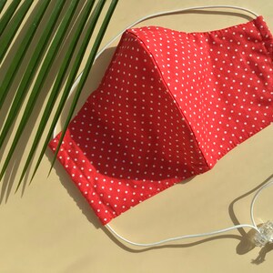 Può includere: Maschera facciale rossa con pois bianchi e lacci elastici bianchi. La maschera è appoggiata su una superficie beige.