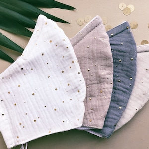 May include: A collection of four face masks in various colors: white, tan, blue-gray, and light beige. Each mask is adorned with small, gold-colored dots. The masks are made of a soft, textured fabric and are arranged in a row, showcasing their design and color variations.