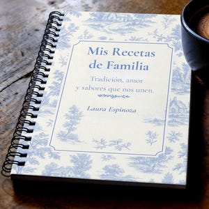 May include: A spiral-bound recipe journal with a blue and white toile pattern. The cover features the text "Mis Recetas de Familia" in a decorative frame. The journal includes the text "Tradición, amor y sabores que nos unen" and the name "Laura Espinoza".