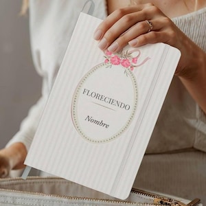 May include: A cream-colored notebook with vertical stripes and a floral design. The cover features an oval frame with the word "FLORECIENDO" and "Nombre" in a delicate font. A pink ribbon and flowers adorn the top of the frame.