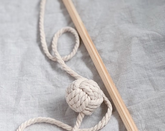 Puede incluir: Un juguete de cuerda de algodón blanco con una bola anudada unida a un palo de madera. El juguete está sobre una superficie de tela gris claro.