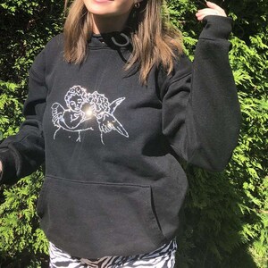 Pu&ograve; includere: Felpa con cappuccio nera con un design di due angeli che si abbracciano in strass. La felpa ha un cappuccio con coulisse e una tasca a marsupio.