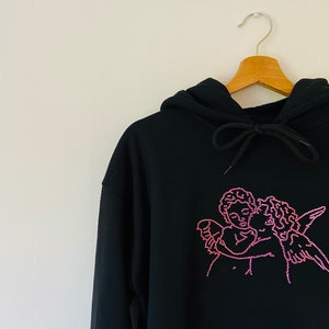 Pu&ograve; includere: Felpa con cappuccio nera con un design di due angeli che si abbracciano in strass rosa.